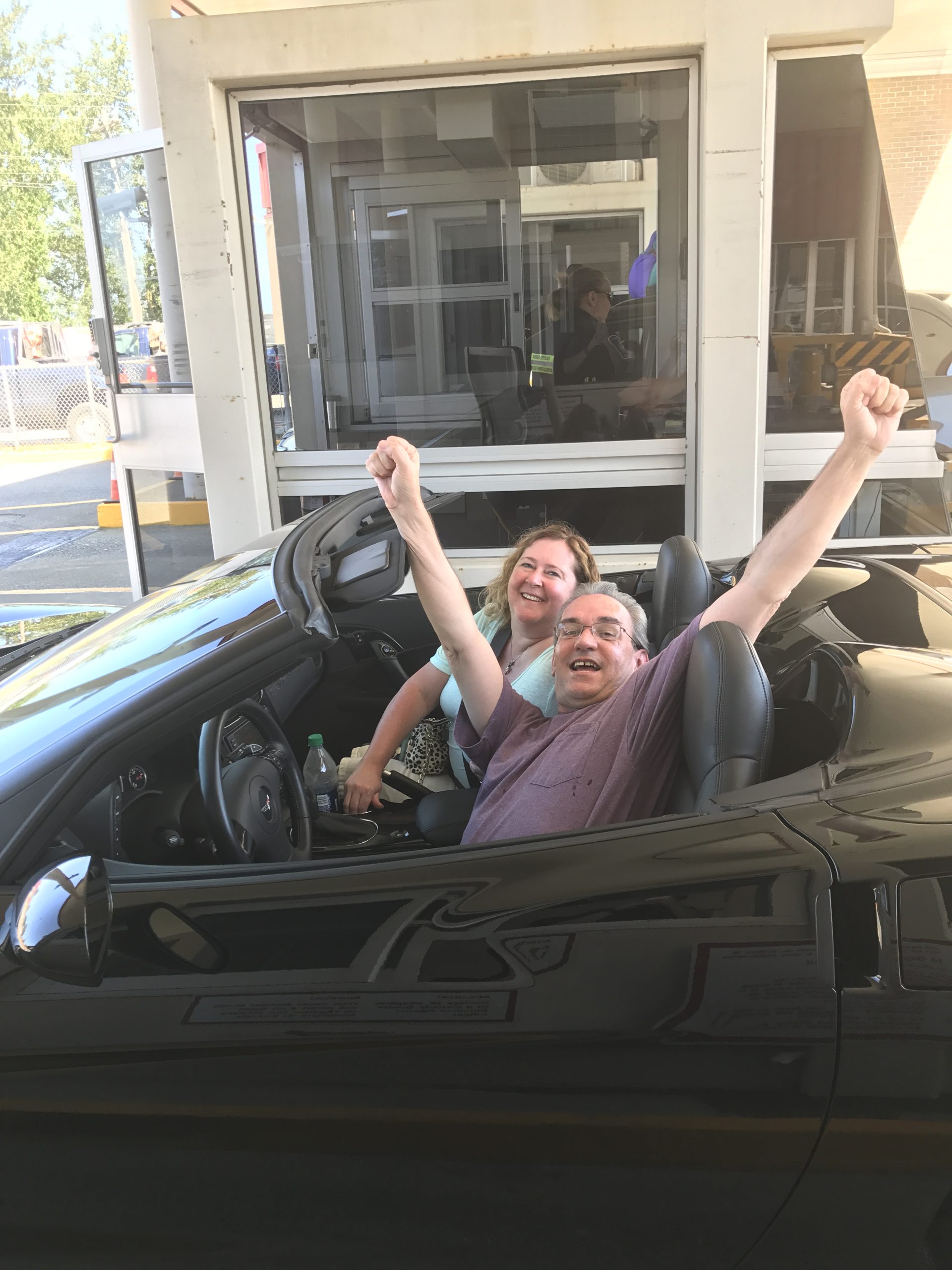 Pam and Bill cheering with their arms raised in their convertible with the top down